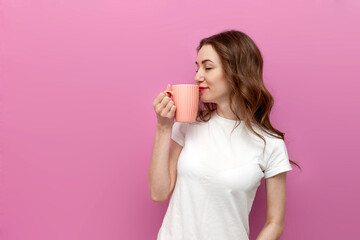 young cute woman in white t-shirt holds cup with drink and smells the aroma on pink isolated background