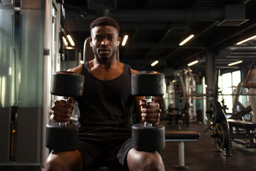 athletic african american man trains in dark gym, athletic guy lifts heavy dumbbells in fitness club - Powered by Adobe