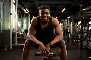 Fototapeta na wymiar young athletic african american man sits in dark gym and smiles, athletic guy rests in fitness club