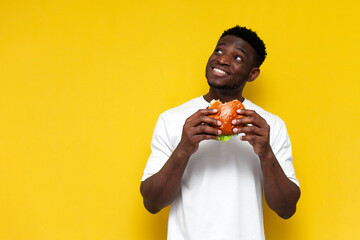pensive african american man in white t-shirt holding big burger and daydreaming, guy eating fast...