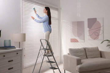 Woman on metal ladder wiping blinds at home © New Africa