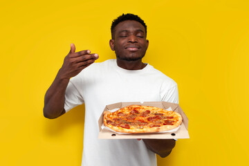 joyful african american man in white t-shirt holding box of pizza and smelling the smell on yellow...