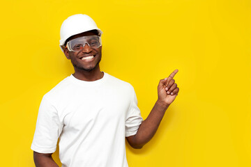joyful male african american builder in uniform smiles and shows his hand to the side on yellow...