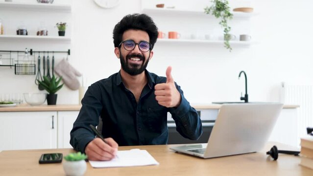 Portrait of attractive indian businessman posing at office desk with digital devices in spacious dining room, thumb up. Self-employed worker carrying out professional activity in comfortable workplace