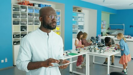 Medium shot of young African American man standing in after school robotics club and using tablet computer, and group of children making models from colourful plastic building blocks in backgroundMedi