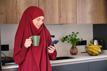Muslim young woman in hijab drinking coffee and using smartphone in the kitchen at home.