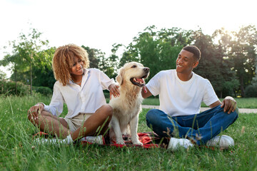 young african american couple with dog sitting in park on green grass, woman and man together with retriever