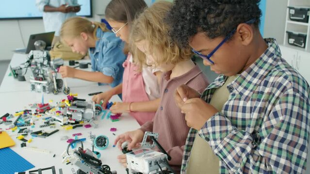 Medium side shot of group of diverse young children in casual clothes standing at table with plastic construction blocks and building electric toy robots, during lesson at after school robotics club