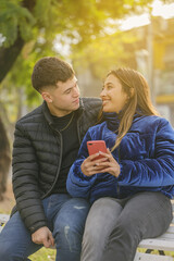 Couple in love sitting on a bench in a public park with mobile phone.