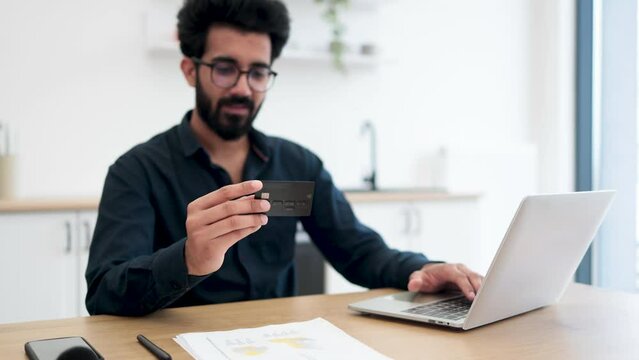 Selective focus of empty black credit card held by cheerful indian man taking seat in cozy workplace at home. Modern remote freelancer checking balances on accounts using online banking services.