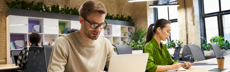 Young focused caucasian man wearing eyeglasses working on laptop while sitting at desk with his...