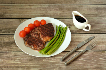 Grilled beef steak, cherry tomatoes and boiled asparagus on a wooden table, next to a gravy boat and cutlery.