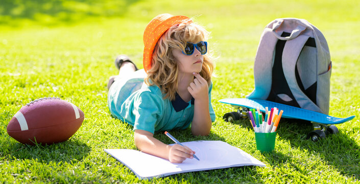 Clever school boy doing homework, writing on copy book in green grass of park.