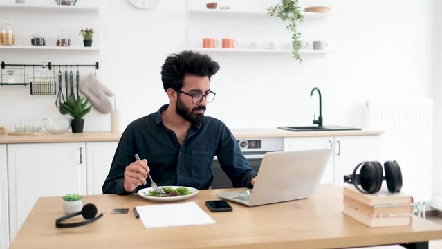 Efficient indian worker in glasses eating vegetables while typing on portable computer at wooden desk in kitchen. Remote freelancer performing business task without wasting time for lunch break.