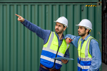 Young shipment worker pointing at container storage location while holding clipboard, explain to colleague about planning for next shipment. A large steel cargo containers is in the background.