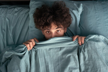 Handsome surprised guy covering half of face with blanket, scared man peeking from duvet, afraid of...