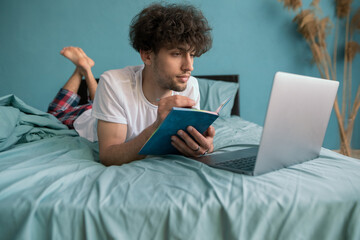 Young arab guy student studying virtual course, learning distance class taking web training, looking at laptop computer, writing notes sitting on bed at home.
