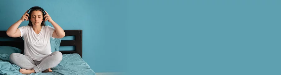 Fotobehang Lotusbloem Young woman meditating and listening to music while sitting on bed at home in lotus position  © Andrii Lysenko