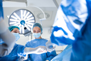 Surgeons operating below lighting equipment. Male and female doctors are wearing blue scrubs. They...