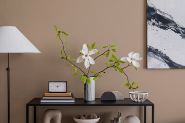 Warm and cozy living room interior with mock up poster frame, black rack, vase with magnolia, books, stylish lamp, clock, sculpture, beige bowl and personal accessories. Home decor. Template.