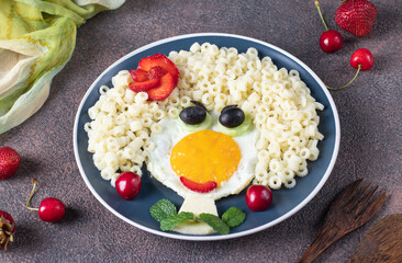 Fun Food for kids - smiling face girl from fried eggs, pasta ditalini, olives, strawberry and cucumber on blue plate