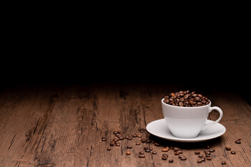 Coffee beans in a white coffee cup and many coffee beans placed around on a wooden table in a warm, light atmosphere, on a dark background, with copy space.