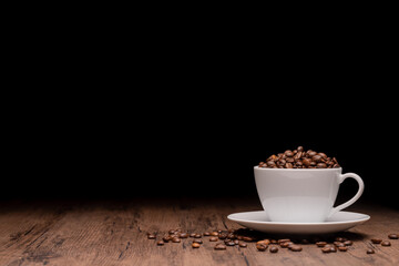 Coffee beans in a white coffee cup and many coffee beans placed around on a wooden table in a warm, light atmosphere, on a dark background, with copy space.