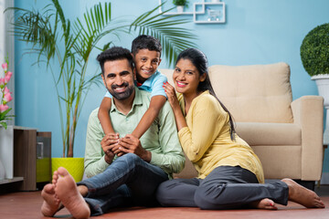 Obraz premium Happy indian parents with son by looking at camera while sitting on floor at home - concept of family bonding, weekend holiday and parenthood.