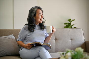 A happy retired Asian woman is daydreaming while relaxing on a sofa in her living room.