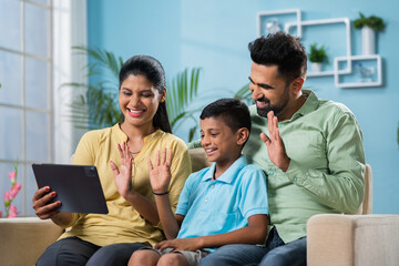 Happy indian family making video call on digital tablet while sitting on sofa at home - concept of distant relationship, and technology