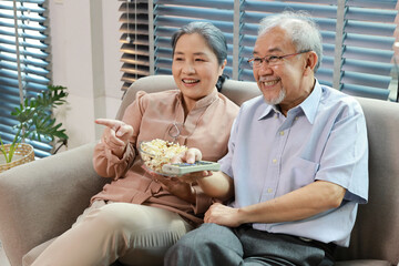 Obraz na płótnie Canvas Happy smiling asian senior man and woman sitting on sofa and eating popcorn while having fun with movie rest indoor at home living room. Couple elder husband and wife embrace are happy while watch TV