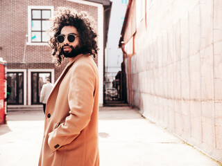 Handsome hipster model.  Unshaven Arabian man dressed in brown coat clothes. Fashion male with long curly hairstyle posing outdoors in street. Cheerful and happy. At sunny day