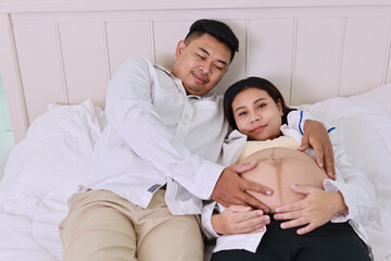 Happy asian adult couple with husband sitting and resting on bed in bedroom while holding baby inside a pregnant and smile. Expectant mother preparing and waiting for baby birth during pregnancy.