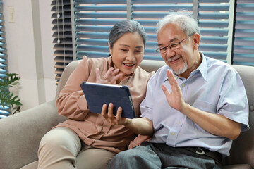 Fototapeta na wymiar Happy smiling asian senior couple sitting on sofa and using tablet while online video call with friend or relative cousin at home living room. Internet information technology and lifestyle concept.