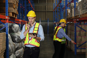 Fototapeta premium Group of warehouse workers with hardhats and reflective jackets using tablet, walkie talkie radio and cardboard while controlling stock and inventory in retail warehouse logistics, distribution center