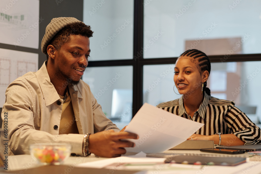 Aufkleber young smiling businessman with papers showing and explaining data to female colleague sitting by des