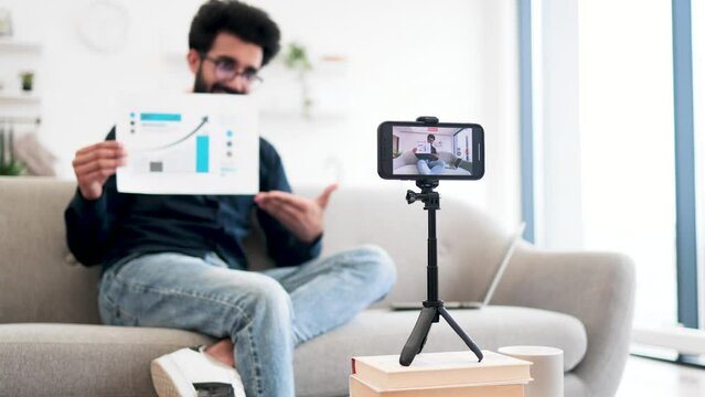 Handsome indian guy in casual clothes sitting on couch with graph financial report in hands while recording video on modern smartphone. Successful financier talking about statistics on camera.