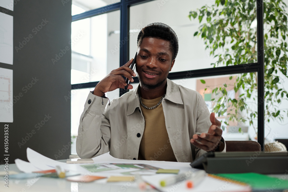 Aufkleber Young African American designer or architect talking on mobile phone while sitting by workplace in office and getting ideas for new project
