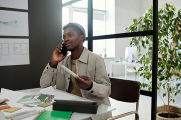 Young confident black man in casualwear talking to colleague on mobile phone while sitting by desk with sketches, catalog and office supplies