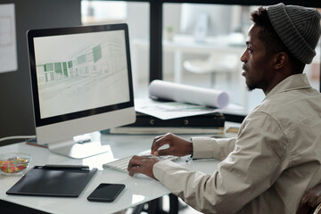 Young black man in casualwear looking at graphic sketches on computer screen while creating drawings for new architectural project