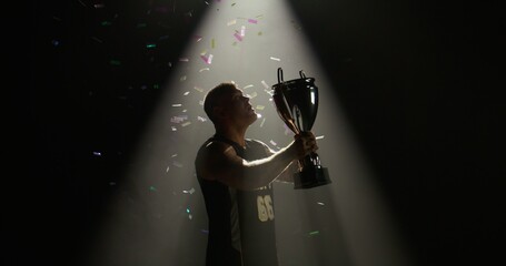 Silhouette of Caucasian male basketball player raising a trophy above head against bright light and falling confetti. Super slow motion, shot on RED cinema camera