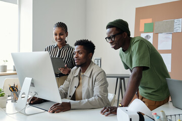 Three young African American graphic designers looking at computer screen at working meeting while one of them drawing sketch