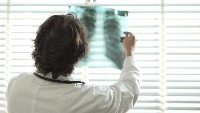 Doctor in room and radiologist discuss diagnosis while watching procedure and monitors showing chest rib bone scans results. Background of patient undergoes MRI or CT scan procedure.