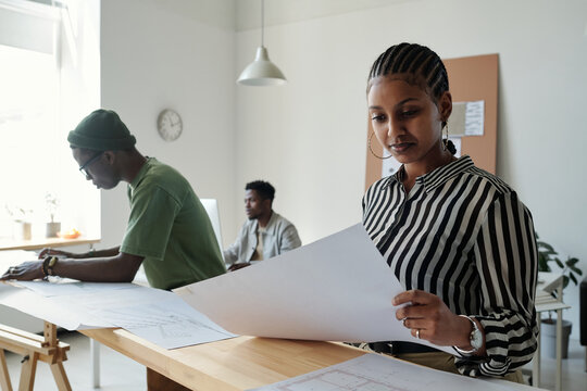 Young serious female architect looking at paper with sketch while sitting by workplace against male colleague bending over blueprint