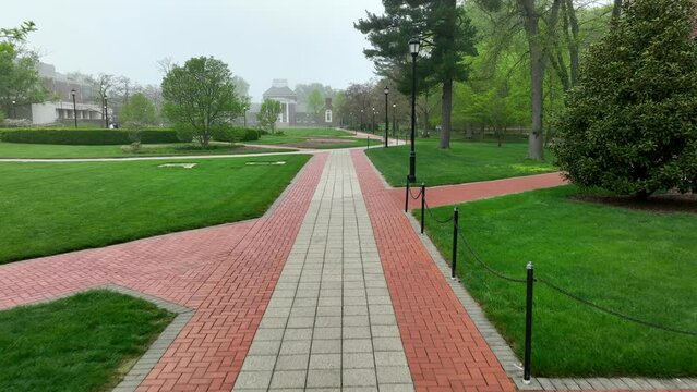 First person aerial shot of brick sidewalks on foggy college campus. Green lawns and historic academic buildings surround green landscaping on walkable University of Delaware campus.