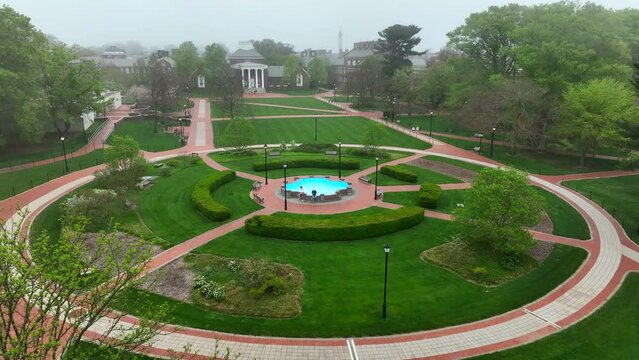 Rotational aerial shot of gardens and fountain in front of manicured lawns and historic academic architecture. Foggy skies over college campus at University of Delaware.