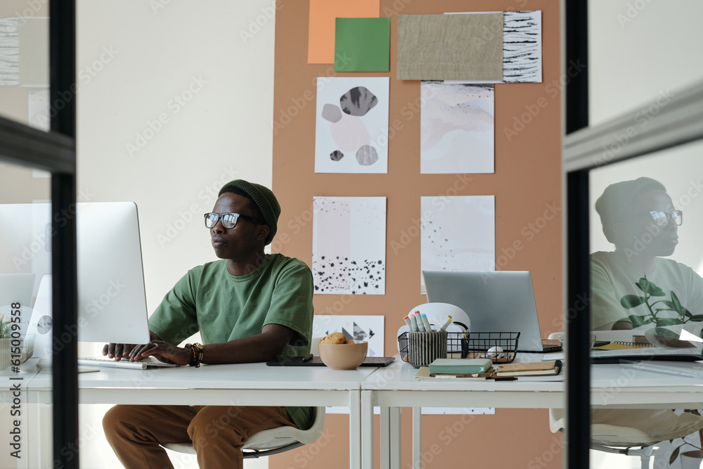 Aufkleber Young creative designer in casualwear and eyeglasses looking at computer screen and typing on keyboard while creating new website in office