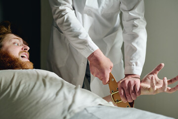 Cropped shot of psychiatrist binding hands of male patient with tight brown belts to calm him down during mental treatment in hospital