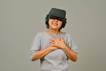 Happy 60s senior woman wearing virtual reality glasses isolated on gray background. Modern technologies and innovations