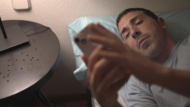 Young Boy Connecting His Phone To Charge Before Falling Asleep. Man In Bed Using His Smartphone, Putting It On Charge And Turning Off The Light Of The Lamp.